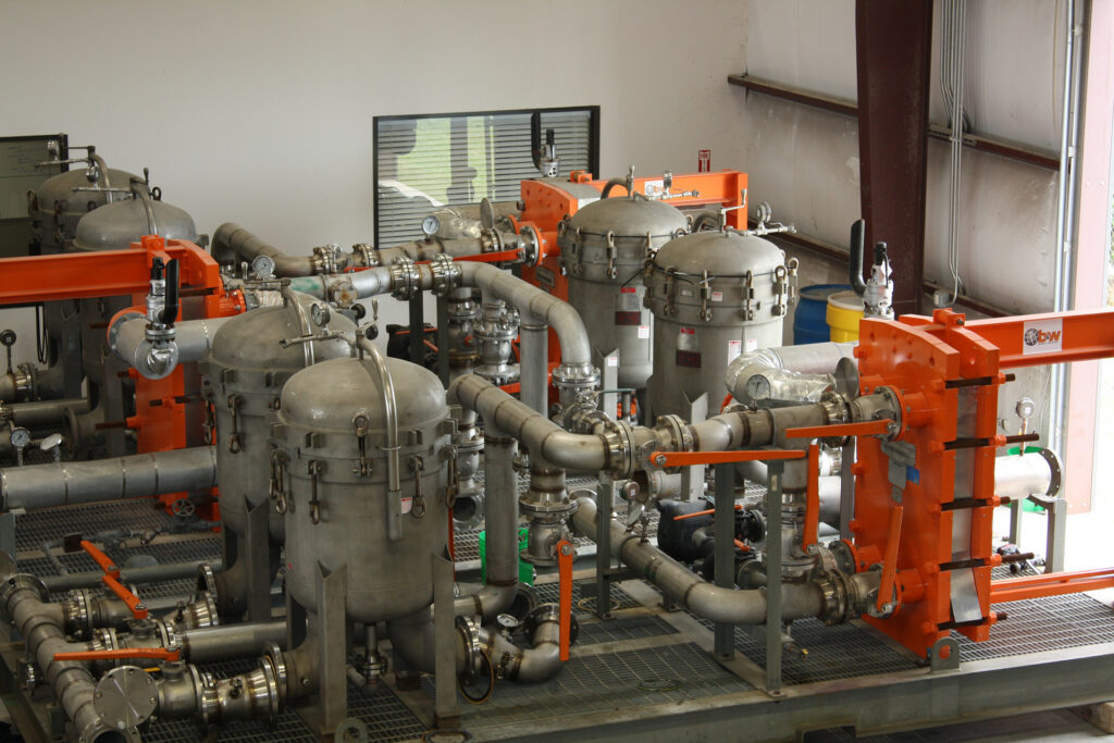 Industrial setup with large gray metal vessels and interconnected pipes on a metal platform. Bright orange frames provide structural support.