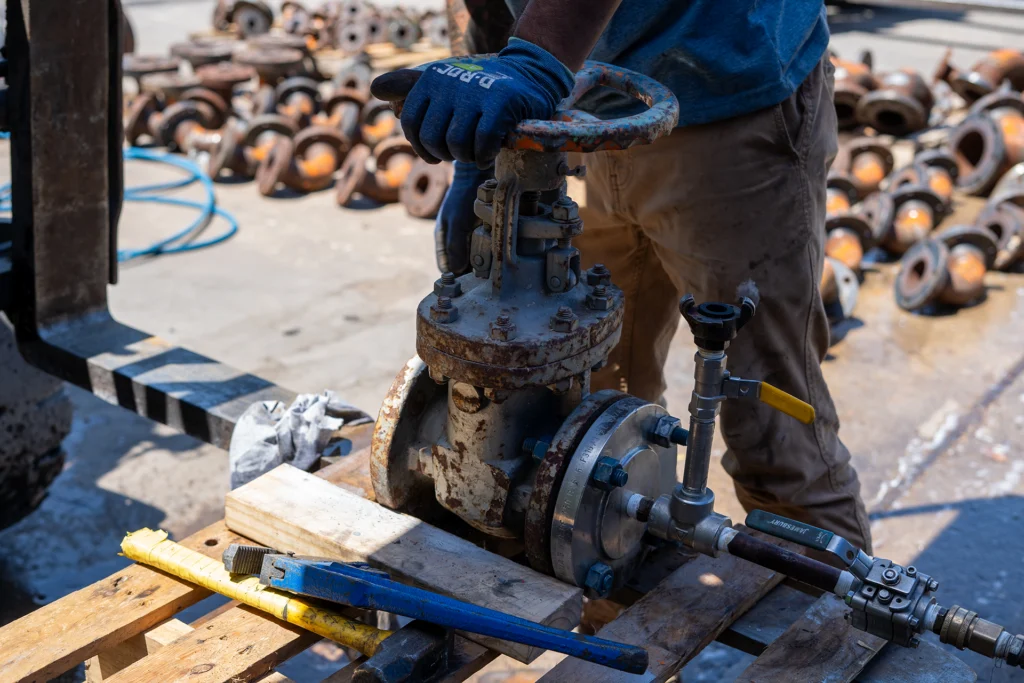 A worker in blue gloves adjusts a large rusted valve on a wooden pallet, surrounded by metal parts on the ground in an equipment yard.