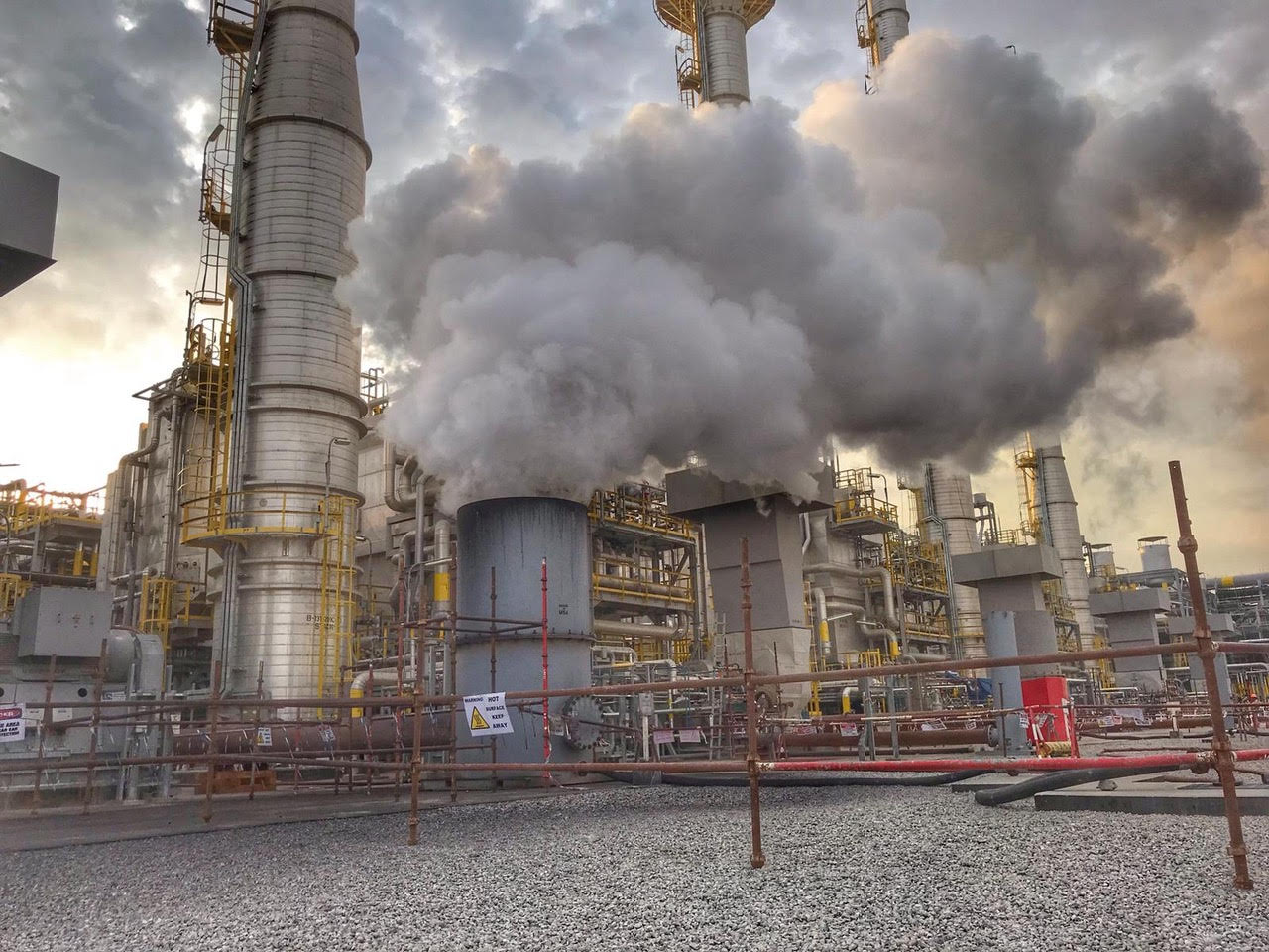 Industrial scene with large metal towers emitting thick white steam, set against a cloudy sky.