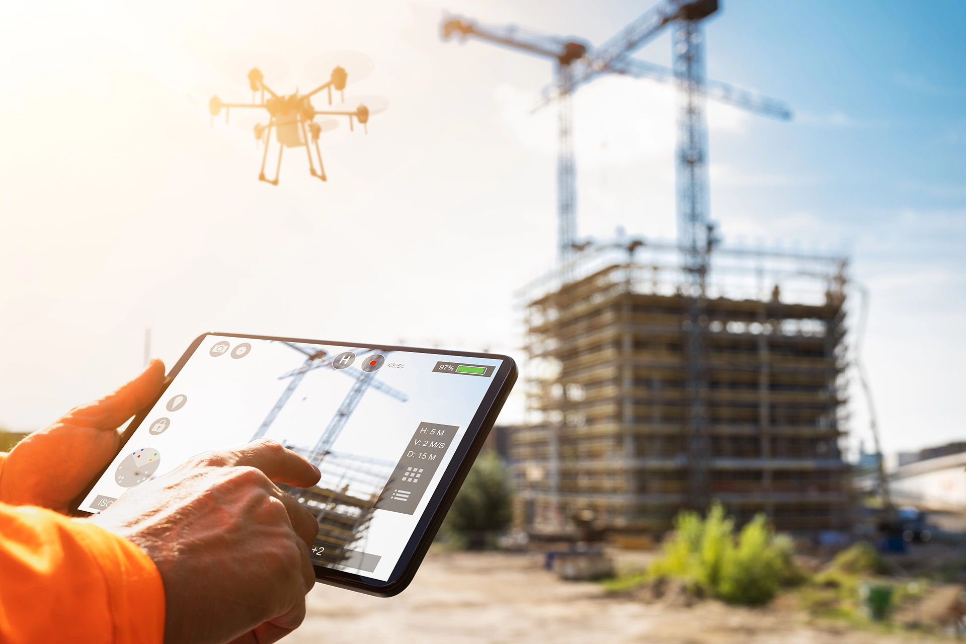 A person operates a drone with a tablet at a construction site. The drone flies near a building under construction, with cranes in the background, under a clear sky.